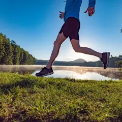 Biegacz w skoku nad jeziorem o poranku, budujący swój VO2 max. Słońce przebija się przez mgłę, a trawa jest wilgotna.