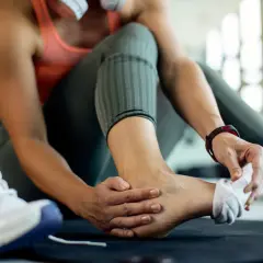 Kobieta w siłowni dotyka bolącej kostki, podejrzewając naderwane ścięgno. Obok leżą buty sportowe.