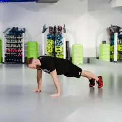 Mężczyzna wykonuje pompki na siłowni, w tle stojaki ze sprzętem.