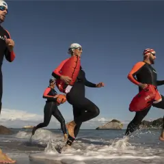 Trzy osoby w piankach biegają po plaży z pomarańczowymi bojami.