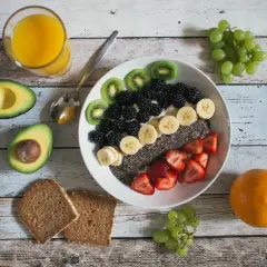 Kolorowa miska pełna owoców: kiwi, jeżyny, banany, truskawki i nasiona chia. Obok awokado, pomarańcza, winogrona i zdrowe śniadanie z pełnoziarnistego chleba.