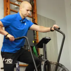 Mężczyzna w niebieskiej koszulce wykonuje trening aerobowy na rowerze stacjonarnym.