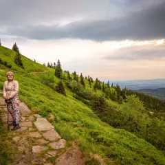 Kobieta z kijkami na kamienistej ścieżce, podziwia widok na zielone zbocze i góry. Widać, ile się wchodzi na Śnieżkę.