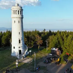 Wieża widokowa na szczycie góry, otoczona lasem. W dole mały domek i ławki, gdzie odpoczywają turyści. Wygląda jak wielka sowa czuwająca nad okolicą.