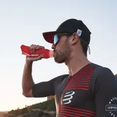 Dżokejka w sportowym stroju pije wodę z czerwonej butelki na tle zachodzącego słońca.