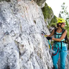 Kobieta w kasku i sprzęcie wspinaczkowym pokonuje via ferraty, zabezpieczona liną przy skalnej ścianie.