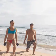 Kobieta i mężczyzna ćwiczą na plaży z hantlami. Ich ruchy to przykład świadomej kinezjologii, poprawiającej siłę i koordynację.