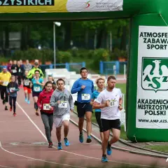 Uczestnicy biegną pod zieloną bramą z napisem "Akademickie Mistrzostwa Polski". Wszyscy biorą udział w teście coopera, pokazując ducha sportowej rywalizacji.