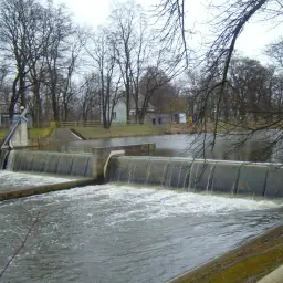 Wodospad na rzece, gdzie woda spływa kaskadami. W tle drzewa i budynki, tworzące spokojny, jesienny krajobraz.