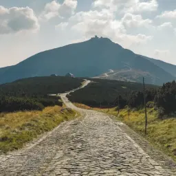 Kamienista droga prowadzi ku górze Śnieżka, otoczonej przez zielone zbocza i chmury.