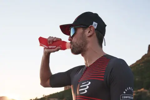 Dżokejka w sportowym stroju pije wodę z czerwonej butelki na tle zachodzącego słońca.