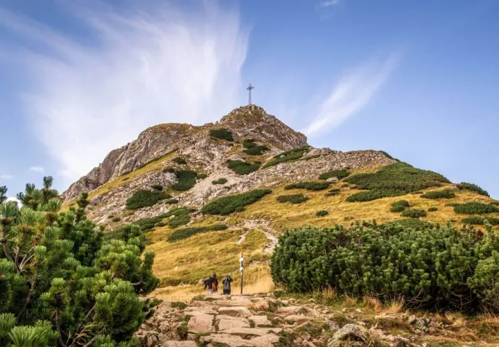 Kamienista ścieżka prowadzi pod górę, gdzie widać krzyż na szczycie. Turyści idą w stronę, by zobaczyć, ile ma Giewont.