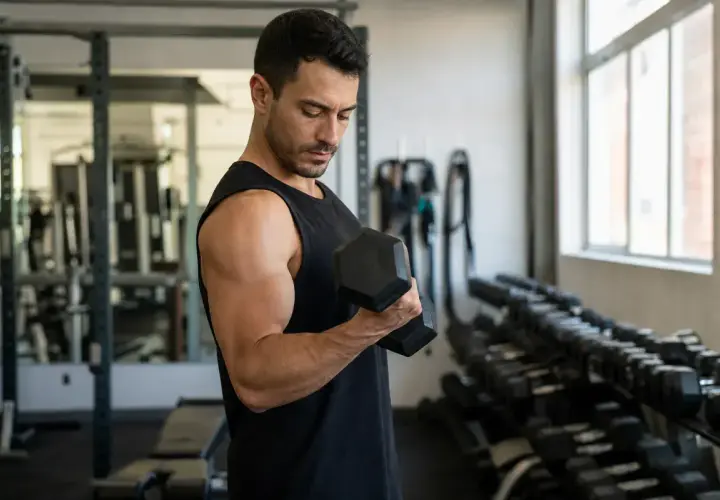 Mężczyzna wykonuje ćwiczenia na biceps z hantlem w siłowni. W tle stojaki z ciężarami.