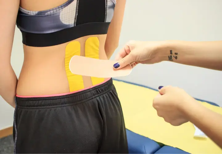 Naklejanie tejpy na plecy w odcinku lędźwiowym. Dłoń z tatuażem "You can do it" przygotowuje plaster.