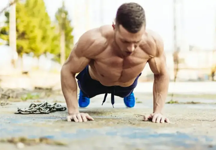 Mięśniowy mężczyzna wykonuje pompki na biceps na zewnątrz. Obok leży ciężki łańcuch.