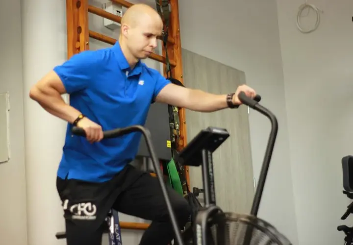 Mężczyzna w niebieskiej koszulce wykonuje trening aerobowy na rowerze stacjonarnym.