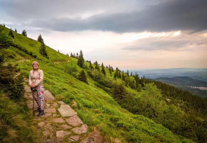Kobieta z kijkami na kamienistej ścieżce, podziwia widok na zielone zbocze i góry. Widać, ile się wchodzi na Śnieżkę.