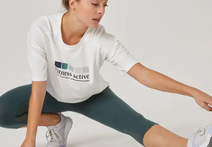 Kobieta w białej koszulce z napisem "Fitness active" wykonuje rozciąganie po treningu siłowym.