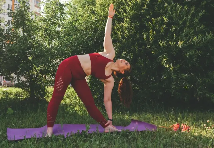 Kobieta w czerwonym stroju wykonuje pozycję Trójkąta podczas praktyki jogi ashtanga na fioletowej macie w trawie.