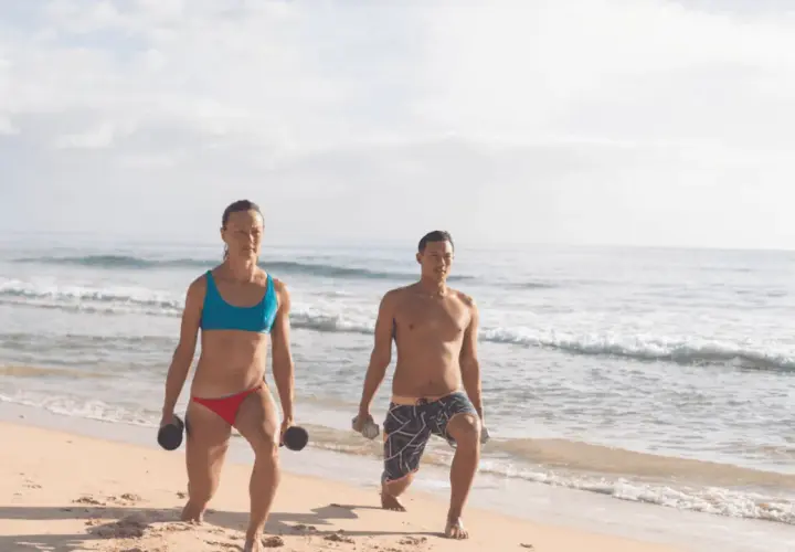Kobieta i mężczyzna ćwiczą na plaży z hantlami. Ich ruchy to przykład świadomej kinezjologii, poprawiającej siłę i koordynację.