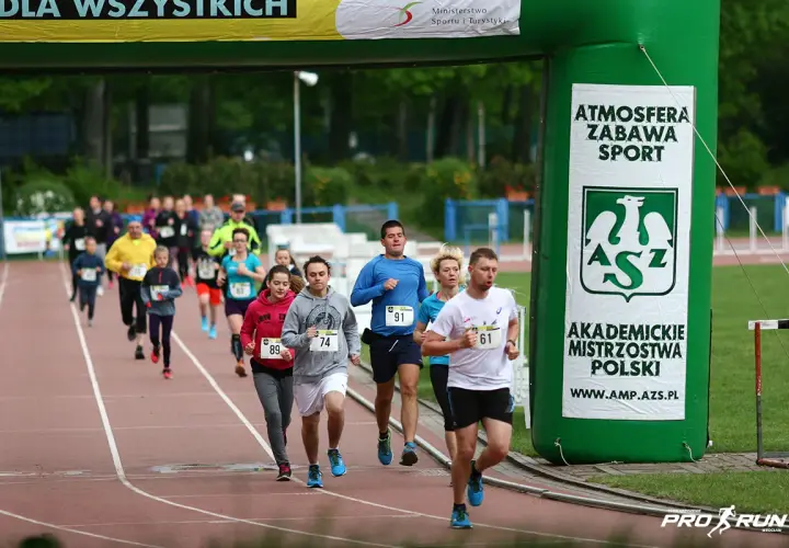 Uczestnicy biegną pod zieloną bramą z napisem "Akademickie Mistrzostwa Polski". Wszyscy biorą udział w teście coopera, pokazując ducha sportowej rywalizacji.