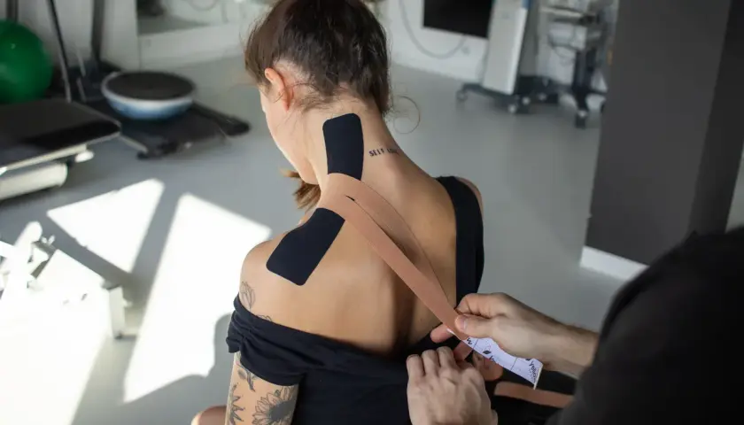 Aplikacja tejpy na szyję i barki dla wsparcia mięśni. W tle sprzęt rehabilitacyjny.