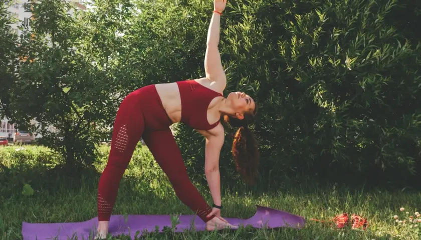 Kobieta w czerwonym stroju wykonuje pozycję Trójkąta podczas praktyki jogi ashtanga na fioletowej macie w trawie.