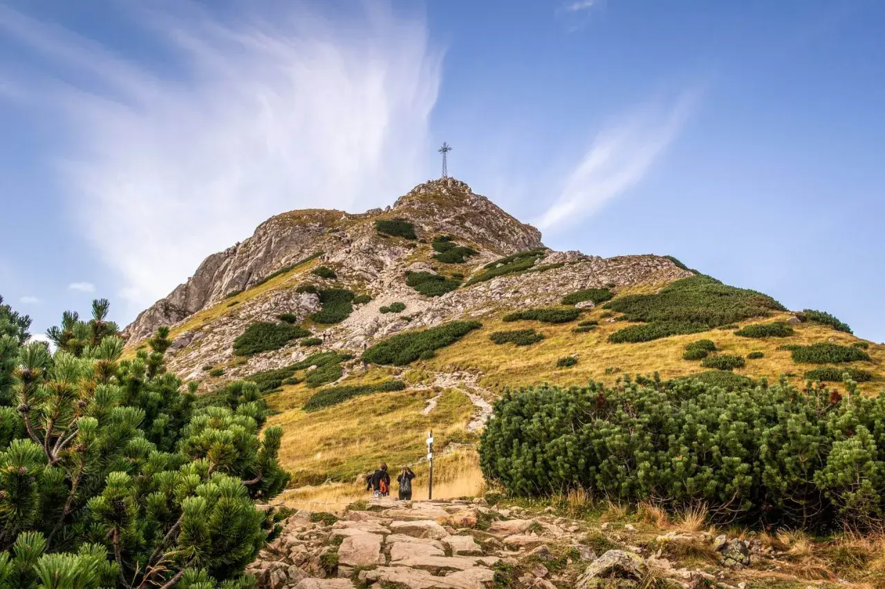 Kamienista ścieżka prowadzi pod górę, gdzie widać krzyż na szczycie. Turyści idą w stronę, by zobaczyć, ile ma Giewont.