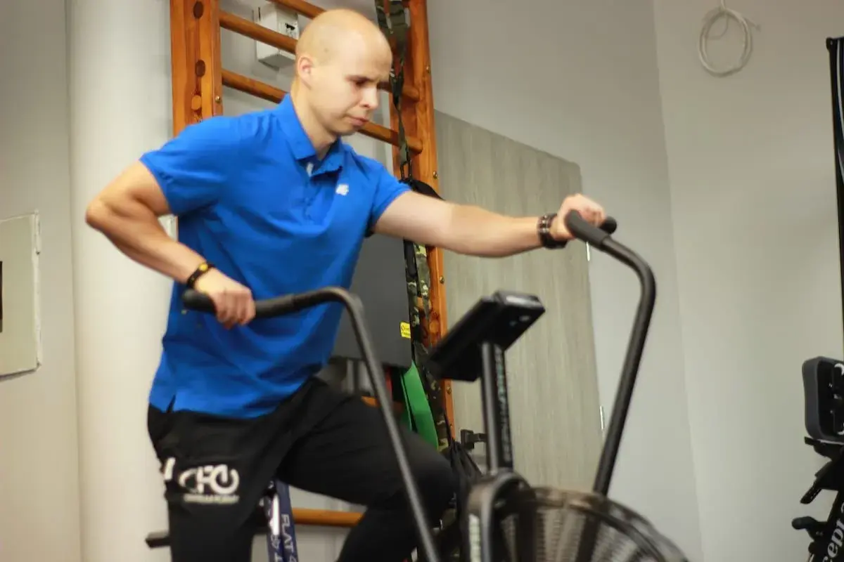 Mężczyzna w niebieskiej koszulce wykonuje trening aerobowy na rowerze stacjonarnym.