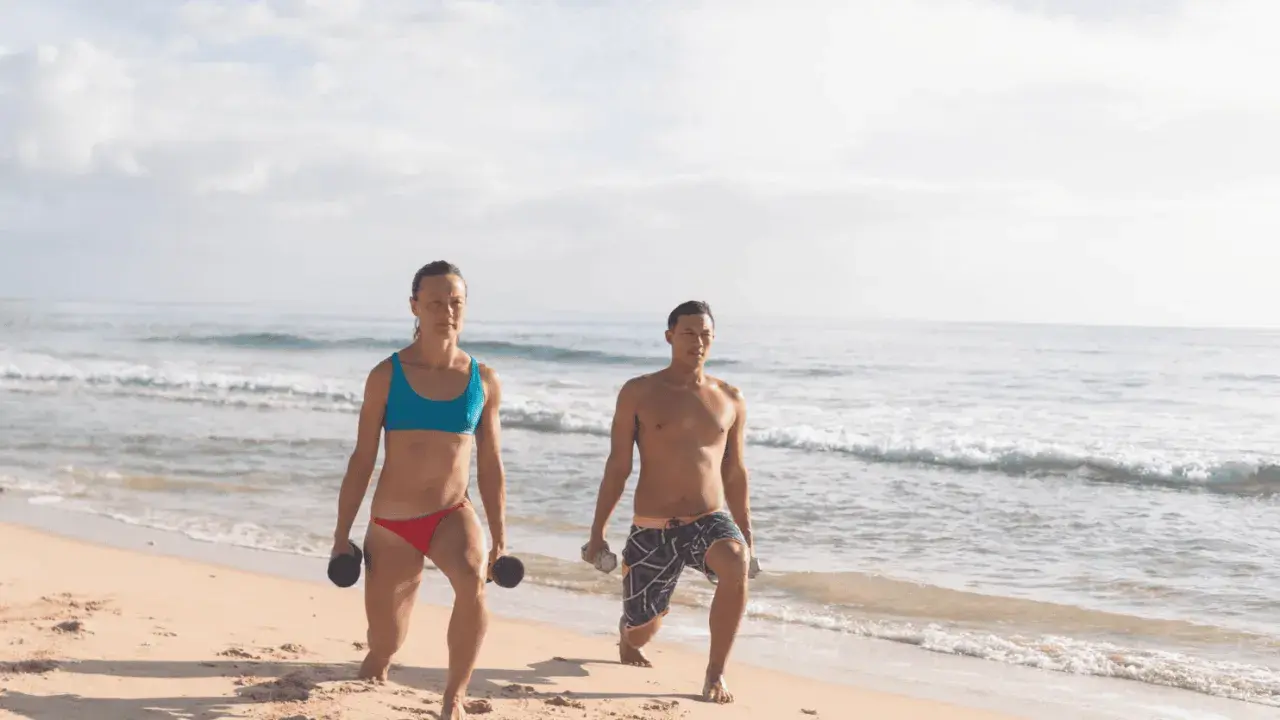 Kobieta i mężczyzna ćwiczą na plaży z hantlami. Ich ruchy to przykład świadomej kinezjologii, poprawiającej siłę i koordynację.