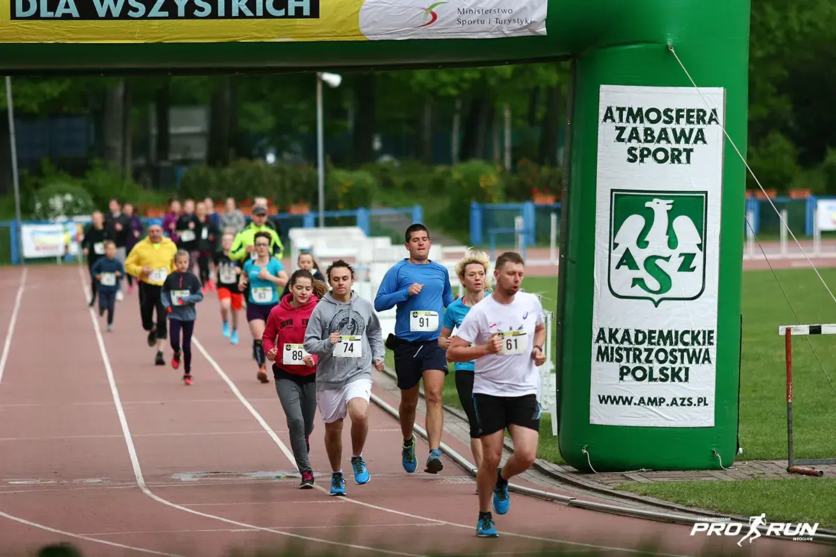 Uczestnicy biegną pod zieloną bramą z napisem "Akademickie Mistrzostwa Polski". Wszyscy biorą udział w teście coopera, pokazując ducha sportowej rywalizacji.