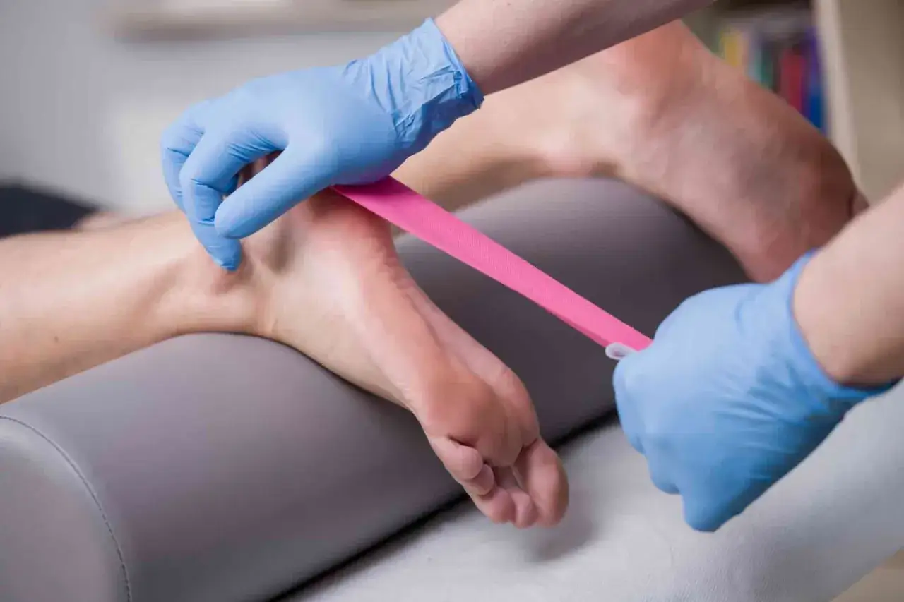Fizjoterapeuta zakłada taśmę kinezjotaping na stopę, wspierając ćwiczenia na ostrogę piętową.