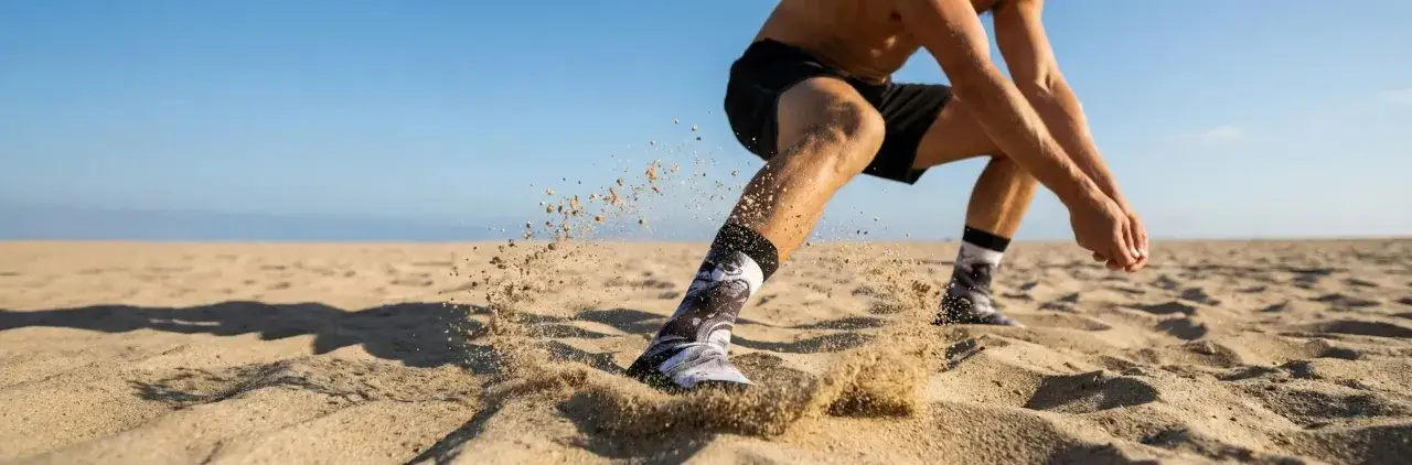 Mężczyzna w sportowych skarpetach wykonuje ćwiczenia na rozgrzewkę na piaszczystej plaży. Piasek wzbija się w powietrze wokół jego stóp.