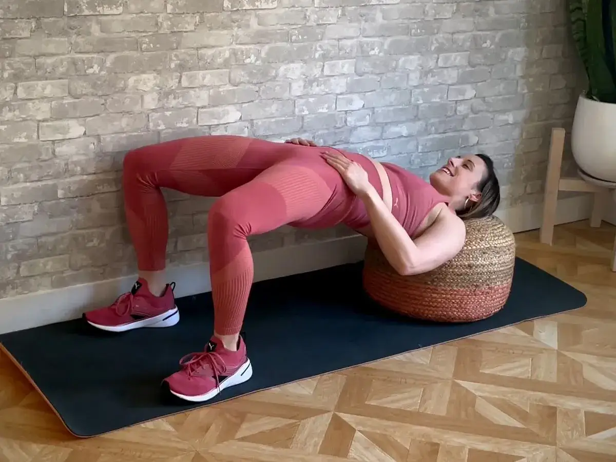 Kobieta wykonuje ćwiczenie hip thrust z nogami opartymi o pufę.