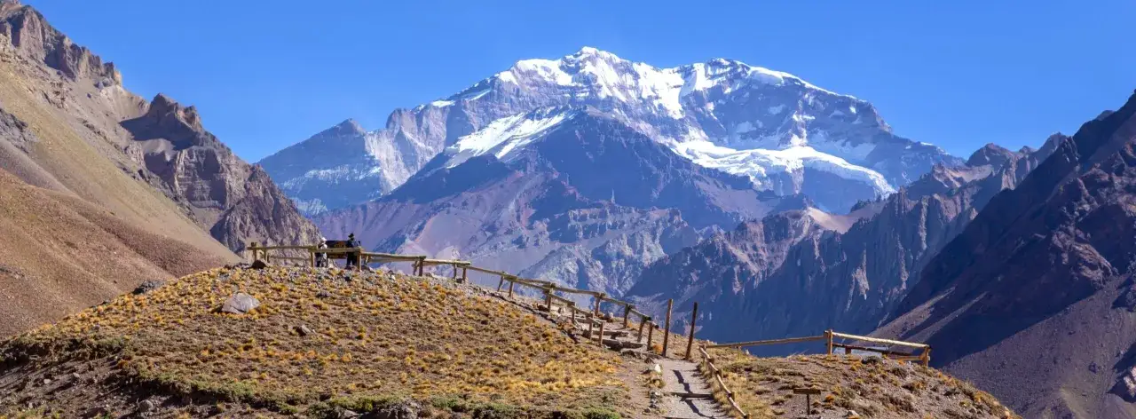 Aconcagua, najwyższy szczyt w Ameryce Południowej, góruje nad surowym krajobrazem. Ścieżka z drewnianym ogrodzeniem prowadzi przez suchą trawę.