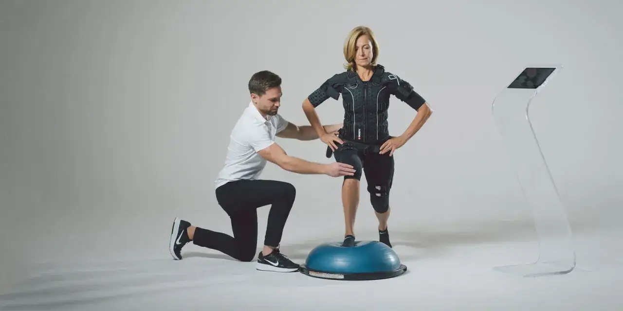 Trener pomaga kobiecie w kamizelce EMS ćwiczyć na piłce BOSU, doskonaląc propriocepcję i równowagę.